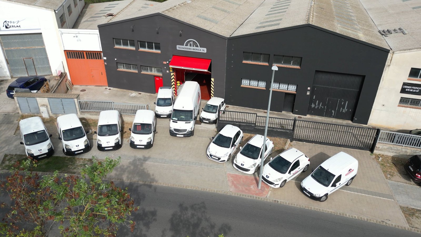 Vista aérea del edificio de Tecnodemo Iberica SL, negro con toldo rojo. Varias furgonetas y coches blancos aparcados en el exterior.