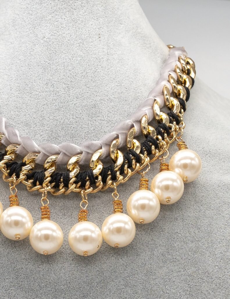 Collar de bisutería con cinta gris trenzada, cadenas doradas entrelazadas y perlas cremas colgantes.